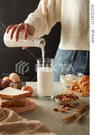 Female hand is pouring fresh milk into a glass decorated with some sandwiches, eggs, nuts, a bowl of flour and a cake on kitchen background. Advertising photo for cooking concept 113932259