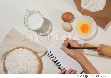 Culinary concept photo with a female hand holding a pencil to write recipe on a notebook, surrounded by some baking pastry ingredients. Space for design or displaying elements Culinary concept photo with a female hand holding a pencil to write recipe on a notebook, surrounded by some baking pastry ingredients. Space for design or displaying elements 113932270