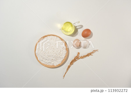 A round wooden dish with flour sprinkled on surface with some eggs, a cup of oil and a wheat branch besides. Minimalist cooking style with blank space for design 113932272