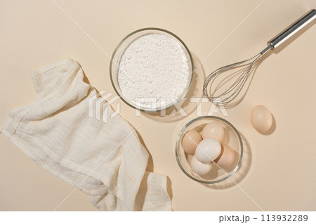 An array of baking ingredients and utensils neatly arranged on pink counter top, including flour, eggs, and whisk, preparation for cooking. Top view photo, space for adding design elements 113932289