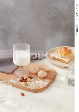 A glasses filled with milk placed on wooden tray, decorated by egg, nuts and gray towel next to a round dish with some cake above and a glass of water. Copy space and blank space for designing A glasses filled with milk placed on wooden tray, decorated by egg, nuts and gray towel next to a round dish with some cake above and a glass of water. Copy space and blank space for designing 113932300