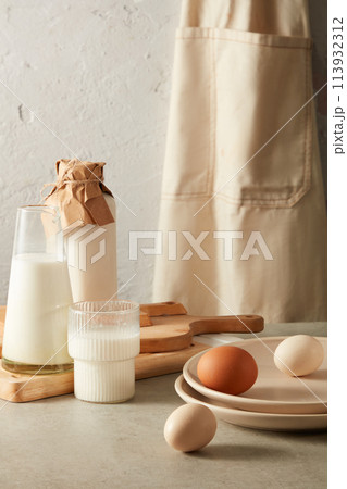 Kitchen essential background with natural milk, eggs, chopping board, cream apron and round plates for displaying dish on white kitchen counter. Empty space for texting and displaying products 113932312