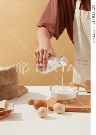 Female hand is holding a milk glass bottle pours milk into a glass bowl next to ingredients for homemade bakery on light brown background. Milk and egg has many benefits for human health 113932329