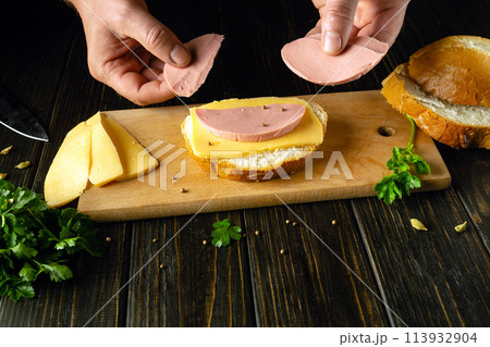 The chef is preparing a sandwich with ingredients. Delicious and fast food on the kitchen table with cheese and sausage with spices The chef is preparing a sandwich with ingredients. Delicious and fast food on the kitchen table with cheese and sausage with spices 113932904