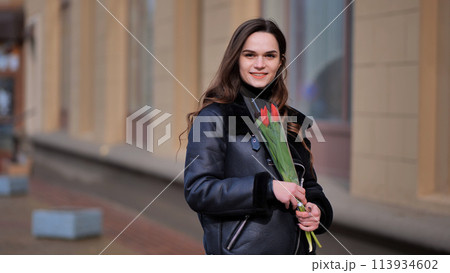 Portrait of a happy brunette girl with a tulip in the background of the city in the cold season. 113934602