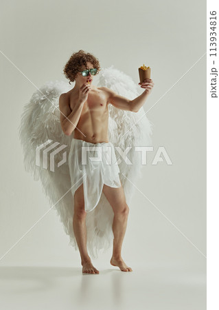 Classical and fast. Yong man with curls an large white wings, looks as angel, hold box of French fries against white studio background. Classical and fast. Yong man with curls an large white wings, looks as angel, hold box of French fries against white studio background. 113934816