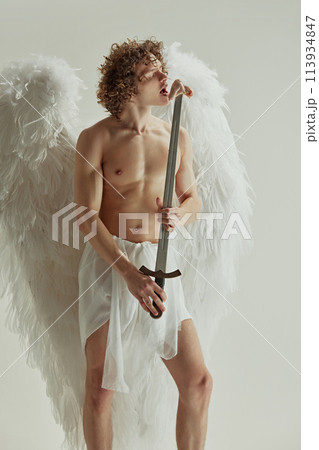 Young man with curly hair and wings poses with sword from which he eats raw meat against white studio background. Young man with curly hair and wings poses with sword from which he eats raw meat against white studio background. 113934847