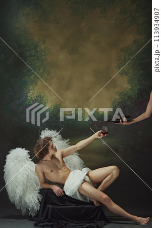 Man with curly hair sits with large wings spread out behind him, reaching out to hand to fill glass with wine against vintage studio background. 113934907