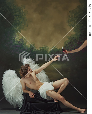 Man with curly hair sits with large wings spread out behind him, reaching out to hand with glass of wine against vintage studio background. Man with curly hair sits with large wings spread out behind him, reaching out to hand with glass of wine against vintage studio background. 113934919