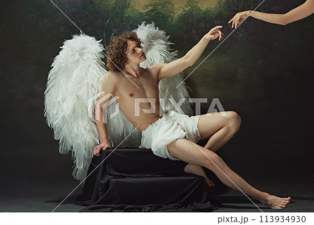 Renaissance painting come to life. Man with curly hair sits with large wings spread out behind him, reaching out to hand against vintage studio background. Renaissance painting come to life. Man with curly hair sits with large wings spread out behind him, reaching out to hand against vintage studio background. 113934930