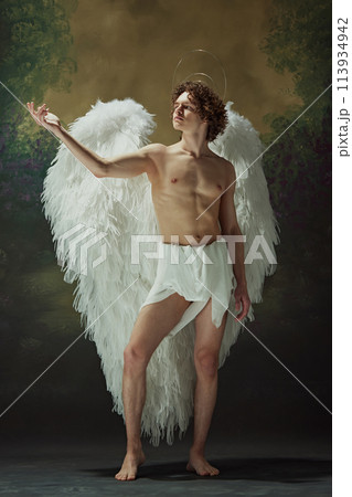 Celestial Elegance. Strikingly handsome man with curly hair and angelic wings poses effortlessly against vintage studio background. 113934942