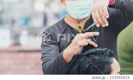 professional man hairdresser wearing face mask holding scissors and haircutting client woman indoor salon, man make business hair care has tool haircut to service haircut to customer, selective focus 113935015
