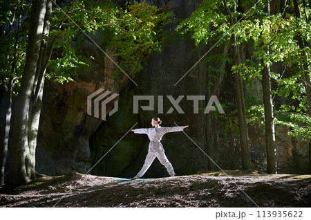 Woman practicing yoga outdoors in forest. Barefoot female on yoga mat surrounded by trees and large rocks, which suggests peaceful, natural environment ideal for meditation or yoga practice. 113935622