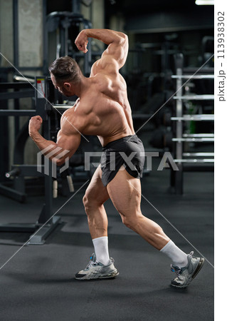 Strong sportsman demonstrating muscular body, while tensing back and arm muscles in gym. Side view of sportive male bodybuilder aesthetically posing with raised hands. Bodybuilding, lifestyle concept. 113938302