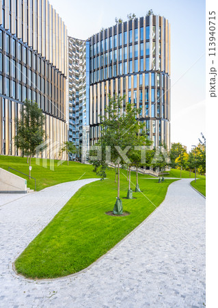 A curving footpath cuts through a lush lawn, leading towards contemporary, curved glass high-rise buildings against a clear sky. 113940715
