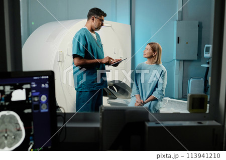 Biracial radiographer holding digital tablet talking to mature Caucasian female patient, through window shot, copy space Biracial radiographer holding digital tablet talking to mature Caucasian female patient, through window shot, copy space 113941210