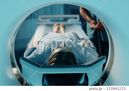 Mature Caucasian woman in blue gown lying on CT scanner bed in modern clinic, unrecognizable doctor adjusting settings 113941215