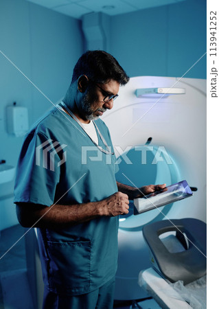 Vertical medium shot of mature biracial doctor using digital tablet at work in modern MRI room, copy space Vertical medium shot of mature biracial doctor using digital tablet at work in modern MRI room, copy space 113941252