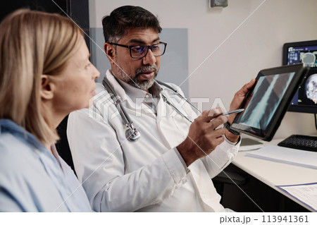 Biracial professional radiographer showing chest X-ray image on digital tablet screen to mature Caucasian female patient, copy space 113941361