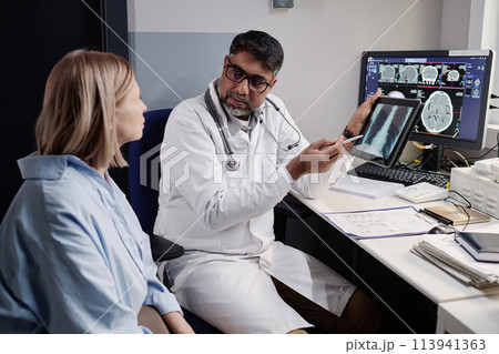 Serious biracial radiographer showing chest X-ray image to mature Caucasian female patient after MRI procedure, copy space 113941363