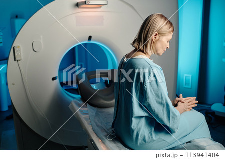 Worried senior Caucasian woman wearing blue patient gown sitting on CT scanner bed in modern hospital, copy space Worried senior Caucasian woman wearing blue patient gown sitting on CT scanner bed in modern hospital, copy space 113941404