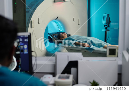 Selective focus shot of mature Caucasian woman having CT scan procedure in modern clinic, unrecognizable doctor looking at computer monitor 113941420