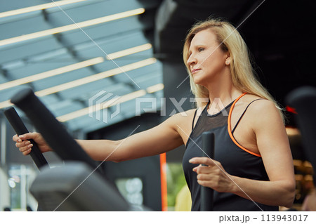 Mature woman in workout gear using an elliptical trainer, focused on her fitness routine at modern gym. Mature woman in workout gear using an elliptical trainer, focused on her fitness routine at modern gym. 113943017