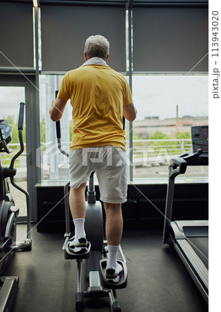 Rear view of senior man in vibrant sports attire with towel on shoulders training on exercise machine modern gym. 113943020