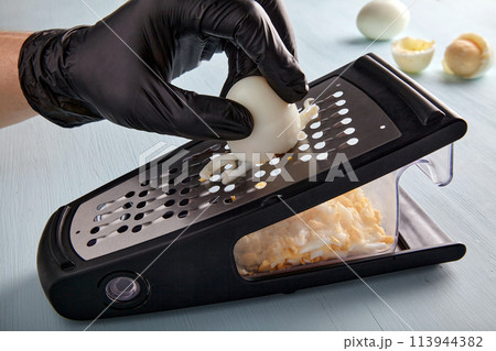 Egg chopped on a grater. Hands in black latex gloves rub a hard boiled chicken egg Egg chopped on a grater. Hands in black latex gloves rub a hard boiled chicken egg 113944382