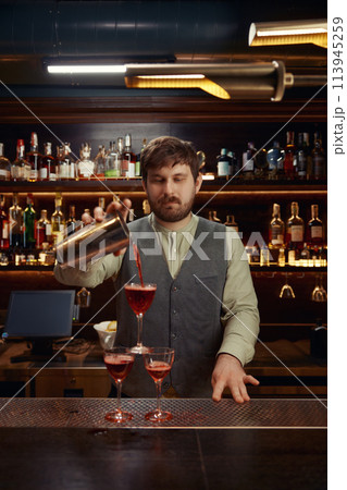 Smiling bartender pouring freshly prepared cocktail into several glasses 113945259