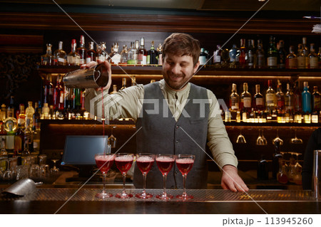 Smiling bartender pouring freshly prepared cocktail into several glasses 113945260
