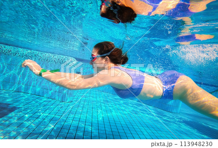 Beautiful girl swimming under water in blue pool wearing swimsuit and googles. Pretty teenager enjoying diving lessons in summer time 113948230