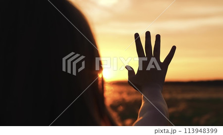 Woman stretched out hand sunset silhouette. Palm arm towards sun catching rays of evening morning dawn light in field meadow. Girl dreamily wistfully reaches for yellow orange sunshine with fingers Woman stretched out hand sunset silhouette. Palm arm towards sun catching rays of evening morning dawn light in field meadow. Girl dreamily wistfully reaches for yellow orange sunshine with fingers 113948399