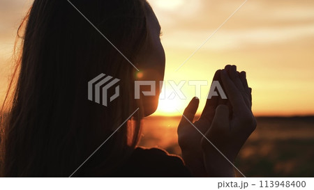 Woman praying with folded hands at sunset silhouette. Religious pious orthodox christian spiritual believing faithful girl at morning dawn sunrise reading prayer bible turning to god jesus christ. 113948400