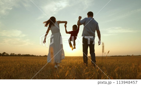Mom dad kid hold hands at sunset. Joyful merry happy family together kid child boy playing jumping parents lifting kid up by hands moving forward strolling across field towards golden setting sun rays Mom dad kid hold hands at sunset. Joyful merry happy family together kid child boy playing jumping parents lifting kid up by hands moving forward strolling across field towards golden setting sun rays 113948457