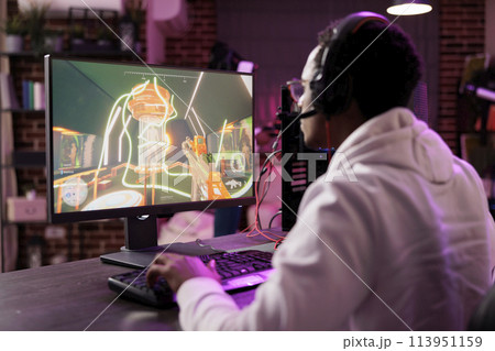 Man in brick wall living room playing video games on gaming PC at computer desk, relaxing after work. Gamer facing enemies in online multiplayer shooter from rgb lights ornate home 113951159