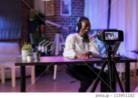 Focus shot on modern camera recording vlogger in blurry background talking about family and relationships subjects. Woman producing online show using high quality equipment in rgb lights studio 113951182