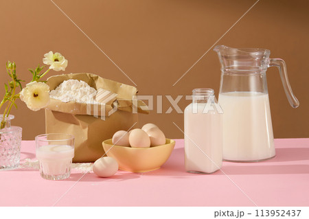 Brown background against cooking concept with milk, eggs and different ingredients decorated by a vase of flower on pink table. Blank space for text or presenting product 113952437