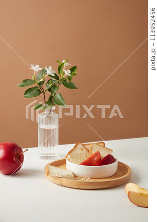 A white bowl on round dish feature some slices of sandwich and apple with a flower vase and an apple on brown background. Front view and blank space for text 113952456