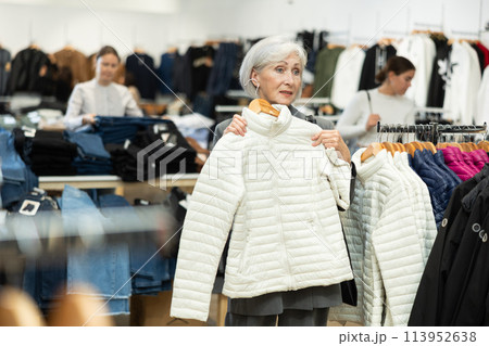 Elderly woman choosing white winter down jacket in clothing shop with large assortment Elderly woman choosing white winter down jacket in clothing shop with large assortment 113952638