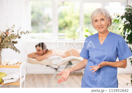 Female elderly physiotherapist poses against background of spa salons office. Female elderly physiotherapist poses against background of spa salons office. 113952682
