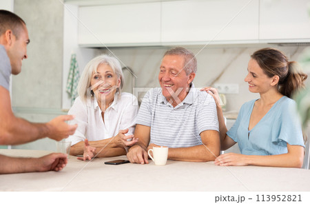 Smiling parents and adult children sit at the table with tea and talk about home life 113952821