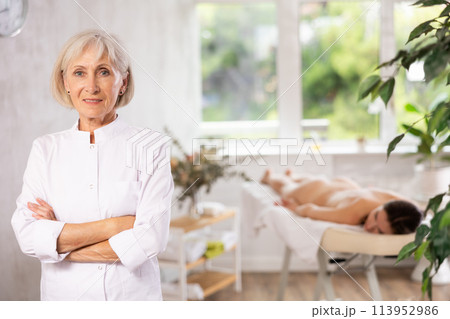 Female elderly physiotherapist poses against background of spa salons office. Female elderly physiotherapist poses against background of spa salons office. 113952986