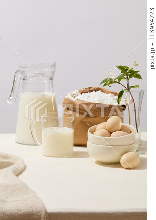 Front view of baking ingredients such as milk, a lot of eggs and a paper bag containing flour. A beautiful pot of flowers decorated. Empty space in the middle for product presentation 113954723