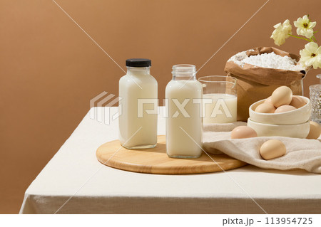 A table with tablecloth features some ingredients for baking such as eggs, flour and milk. A wooden chopping board with two blank label milk bottles displayed on for product promotion 113954725