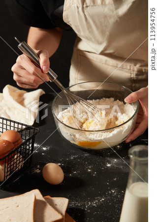 A baker wearing apron is using a whisk to mix eggs and flour inside a glass bowl. Black table featured a basket of eggs, sandwiches and a bottle of milk 113954756