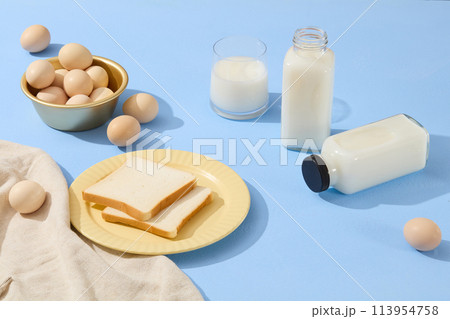 A yellow dish featured two slices of sandwich arranged with a golden bowl containing eggs. Milk is filled inside a cup and bottles. An beige apron is featured on blue surface A yellow dish featured two slices of sandwich arranged with a golden bowl containing eggs. Milk is filled inside a cup and bottles. An beige apron is featured on blue surface 113954758