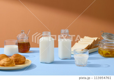 Empty label bottles filled with milk displayed with dishes of bread and sandwiches. A jar of honey, a milk cup and and a jar of jam are arranged on brown background 113954761