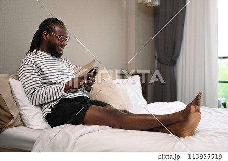 Smiling African guy checking messages on his mobile phone lying in bed at home Smiling African guy checking messages on his mobile phone lying in bed at home 113955519