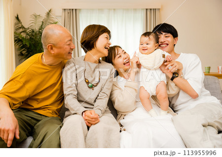 リビングで父親に抱っこされる0歳児の赤ちゃんと母親と祖父母の3世代で複数人の家族 113955996